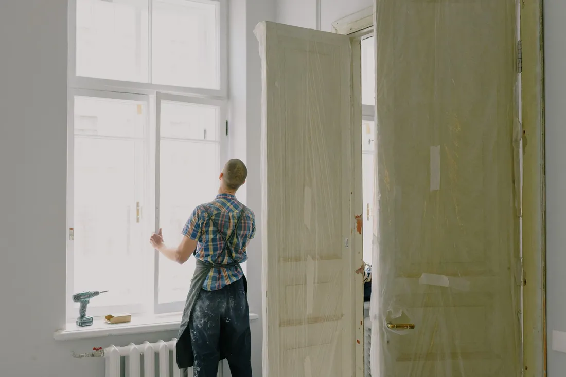 Appartement ancien en rénovation : hautes fenêtres, portes protégées par un film, artisan en tablier