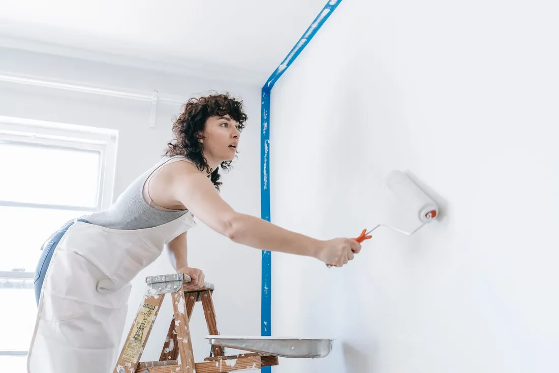 Mur repeint en blanc au rouleau depuis un escabeau, angles protégés par du ruban de masquage